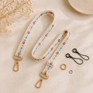 Può includere: Cinturino in tessuto color crema con motivo floreale blu e viola. Il cinturino ha chiusure in metallo color oro e una fibbia regolabile bianca. Sono inclusi anche due anelli neri e due anelli metallici.