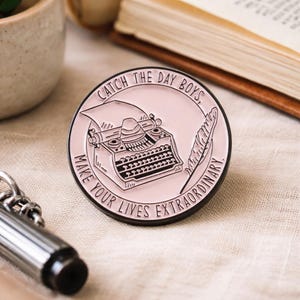 Könnte beinhalten: Runder, rosa Emaille-Pin mit schwarzem Rand. Der Pin zeigt eine alte Schreibmaschine, eine Feder und den Text "CATCH THE DAY BOYS, MAKE YOUR LIVES EXTRAORDINARY" in einem kreisförmigen Design. Ein silbernes Metallobjekt befindet sich im Vordergrund.