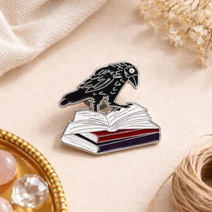 Op de afbeelding: Een zwarte raven pin op een stapel boeken. De pin heeft een zilveren rand en een gedetailleerd ravenontwerp. De boeken zijn wit, rood en paars, met een zilveren rand. Een decoratief accessoire.