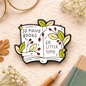 Könnte beinhalten: Weißer Emaille-Anstecker in Form eines offenen Buches mit dem Text "SO MANY BOOKS SO LITTLE TIME". Der Anstecker hat grüne Blätter und rote Beeren mit schwarzer Umrandung. Ein charmantes Accessoire für Bücherliebhaber.