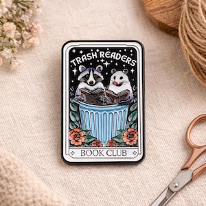 Könnte beinhalten: Rechteckiger Emaille-Pin mit schwarzem Rand und dem Text "Trash Readers Book Club". Der Pin zeigt einen Waschbären und einen Hamster, die Bücher in einer blauen Mülltonne lesen, umgeben von Blumen. Ein einzigartiges Accessoire.
