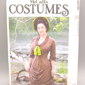 May include: McCall's Costumes sewing pattern featuring a woman in a brown dress with a green bow and hat. The pattern includes sizes A5 (6-8-10-12-14). The pattern is designed by Angela Clayton.