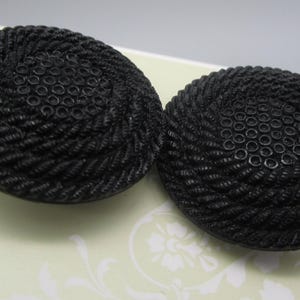 May include: A pair of round, black buttons featuring a textured, rope-like spiral pattern radiating from a central circle of small, raised dots. The buttons have a matte finish and a distinct, molded design that creates a tactile, circular appearance.