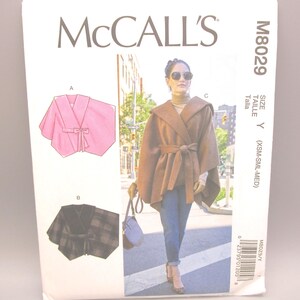 May include: McCall's sewing pattern M8029 featuring a cape design. The pattern includes illustrations of a pink cape with a bow, a plaid cape, and a brown hooded cape. The size is Y (XSM-SML-MED).