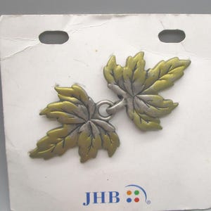 May include: A metal leaf-shaped brooch with a silver and gold finish. The brooch is attached to a white card with two holes at the top. The card has the letters "JHB" and a logo at the bottom.