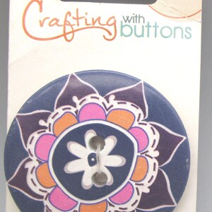 May include: A navy blue wood button with a floral design in pink, orange, and purple. The button has two holes for sewing and is packaged on a white card with the words "Crafting with buttons".