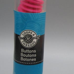 May include: A clear plastic tube containing stacked, bright pink buttons. The tube has a black cap with a loop and a label with the text "Loops & Threads" and "Buttons" in English, French, and Spanish. Each button is approximately 5/8 inch (16 mm) in diameter.