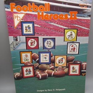 May include: A softcover book titled Football Heroes II Book 2 with a cover featuring a stadium background, multiple framed cross-stitch patterns of football players, and several brown leather footballs. The text reads Designs by Harry D. Hedgepath.