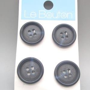 May include: Four dark gray, round buttons with four holes and metal shanks are displayed on a white card. The card has a light blue header with the text "Le Bouton". The buttons are for sewing and craft projects.