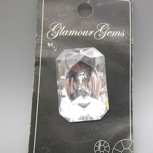 May include: A clear, faceted rectangular ring on a black card with the text "Glamour Gems". The ring has a silver metal band. The card has a white border and a small hole at the top.