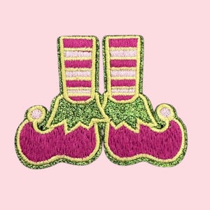 Könnte beinhalten: Gestickter Aufnäher mit einem Paar Elfenstiefeln. Die Stiefel sind magenta mit grünem Glitzersaum und gelb-weiß gestreiften Socken. Der Aufnäher ist vor einem hellrosa Hintergrund platziert.