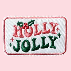 Może przedstawiać: Haftowana naszywka z napisem "HOLLY JOLLY" w kolorze czerwonym i zielonym, z jagodami i liśćmi ostrokrzewu. Naszywka ma białe tło z czerwoną obwódką i ma kształt prostokąta.