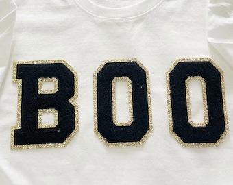 May include: White t-shirt with a black "BOO" graphic made of felt with gold glitter trim.