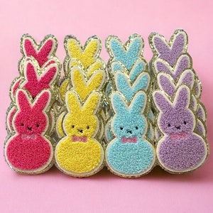 May include: A collection of colorful felt bunny shaped pins with glitter accents. The bunnies are pink, yellow, and blue and have a bow tie.