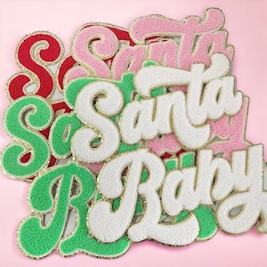 Op de afbeelding: Een stapel kleurrijke viltpatches met de woorden "Santa" en "Baby" in een retro-lettertype. De patches zijn gemaakt van pluche vilt en hebben een gouden glitterrand.
