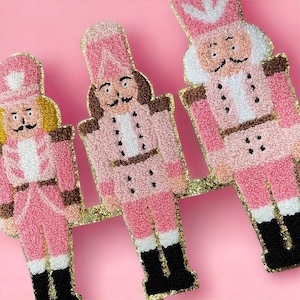 May include: Three pink and white nutcracker figurines with gold glitter accents. The nutcrackers are standing in a row on a pink background.