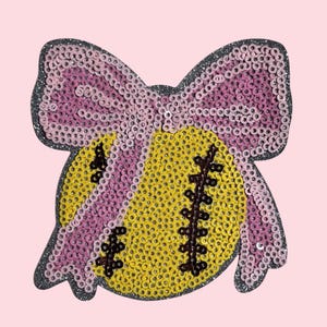 May include: A sequined patch in the shape of a softball with a pink bow. The softball is yellow with black stitching, and the bow is pink. The patch has a glittery gray outline and is set against a light pink background.
