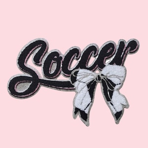 May include: A pink patch with the word "Soccer" in black script with a silver outline and pink accents. A black and white bow is below the word. The patch is likely for clothing or accessories.