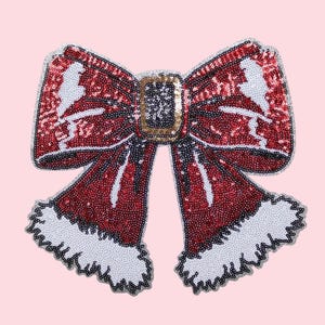 Puede incluir: Un lazo decorativo grande con lentejuelas rojas, detalles blancos y una hebilla dorada. El lazo tiene un tema navideño, con ribete blanco a lo largo de los bordes inferiores. El lazo es probablemente para decoraciones navideñas.