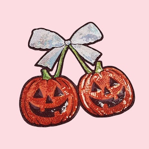 Op de afbeelding: Twee oranje pailletten pompoenen met lachende gezichten, verbonden door groene stelen en bekroond met een zilveren pailletten strik, tegen een lichtroze achtergrond. Een feestelijk Halloween-thema.