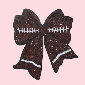 Puede incluir: Un lazo decorativo grande con temática de fútbol americano. El lazo está hecho de lentejuelas marrones con detalles de costura blancos y un borde negro. El lazo está sobre un fondo rosa claro.