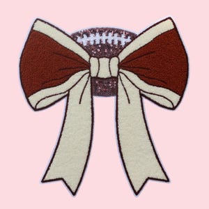 Puede incluir: Un parche grande con forma de lazo, con un centro marrón y bordes y cintas blanquecinas. El centro del lazo es un óvalo de lentejuelas. El parche está sobre un fondo rosa claro, creando un contraste llamativo.