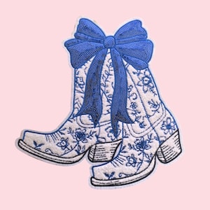 Puede incluir: Parche bordado con un par de botas de vaquero blancas con diseños florales azules y un gran lazo azul. Las botas tienen una apariencia texturizada y un contorno negro, sobre un fondo rosa claro.