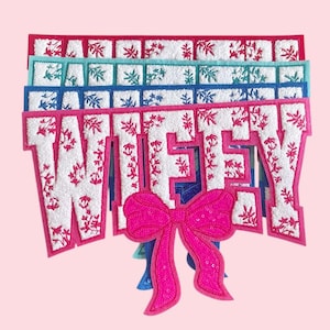 Puede incluir: Un conjunto de parches decorativos que deletrean "WIFEY" en blanco, con detalles florales rosas. Se adjunta un gran lazo rosa con lentejuelas. Los parches están superpuestos en rojo, turquesa, azul y rosa, con patrones florales.
