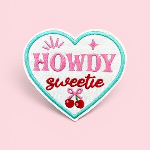 May include: A heart-shaped embroidered patch with a white background and a light blue border. The text HOWDY is in pink, and sweetie is in red. Above the text are pink starburst and diamond icons. Below the text are two red cherries with a pink bow.