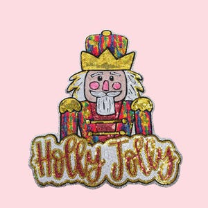 Puede incluir: Un parche de lentejuelas con un colorido soldado cascanueces con una corona y detalles dorados. El parche tiene las palabras "Holly Jolly" en una fuente decorativa. El fondo es de color rosa claro liso.