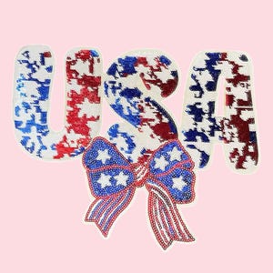 Op de afbeelding: Een patriottisch ontwerp met het woord "USA" in witte letters met rode en blauwe pailletten accenten. Een strik met witte sterren en rood-blauwe strepen staat onderaan. De achtergrond is lichtroze.