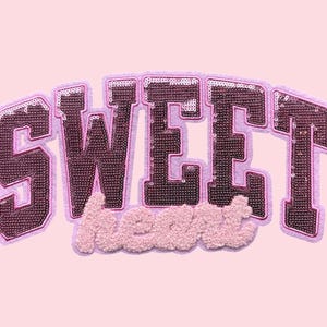 May include: A pink patch with the word "SWEET" in dark pink sequined letters, outlined in light pink. Below, the word "heart" is in a fluffy, light pink material. The background is a solid light pink.