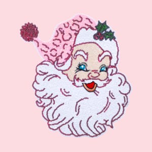 Puede incluir: Parche con la cara de Papá Noel con lentejuelas, con gorro y barba rosas y ojos azules. La cara de Papá Noel sonríe con la boca roja y la barba blanca. El parche está sobre un fondo rosa.