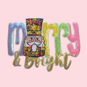 May include: A pink background with a sequined design featuring a colorful nutcracker and the words "Merry & Bright". The text and nutcracker are embellished with sequins in various colors, including blue, green, yellow, and pink.