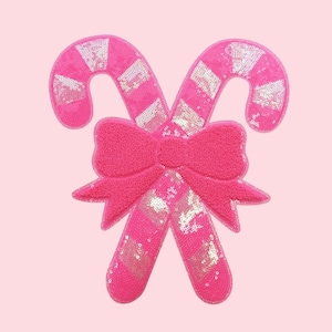 Puede incluir: Dos bastones de caramelo rosas con un lazo en el centro. Los bastones de caramelo están decorados con lentejuelas plateadas en un patrón diagonal. El lazo está hecho de un material rosa texturizado. El fondo es de color rosa liso.