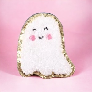 May include: A white fuzzy ghost with a gold glitter outline and a smiling face. The ghost has pink cheeks and black eyes.