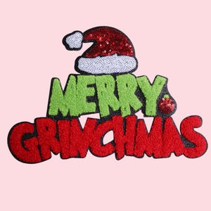 Puede incluir: Parche festivo con las palabras "MERRY GRINCHMAS" en verde y rojo, coronado por un gorro de Papá Noel. Las letras y el gorro están adornados con lentejuelas y una textura difusa, sobre un fondo rosa claro.