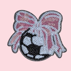 May include: A sequined patch featuring a soccer ball design. The soccer ball is black and white, and is set against a bow made of pink, white, and silver sequins. The patch has a silver glitter border and is set against a pink background.