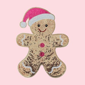 Peut inclure: Ornement de Noël en forme de bonhomme en pain d'épices à paillettes. Le bonhomme en pain d'épices est beige avec des accents marron et des bordures blanches. Il porte un bonnet de Père Noël rose et des boutons roses.