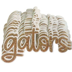 May include: A pile of white and gold sequined "gators" patches. The word "gators" is written in a cursive font, with gold sequins outlining the letters. The patches are white with gold sequin accents.