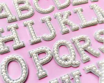 Puede incluir: Primer plano de letras del alfabeto, cada letra es blanca con un borde plateado y adornada con perlas de imitación. Las letras están dispuestas sobre un fondo rosa. Son letras mayúsculas.
