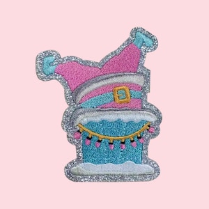 May include: Embroidered patch featuring a whimsical design of a blue and pink elf hat atop a blue cake with a string of pink lights. The patch has a silver glitter border and a gold buckle on the hat.