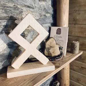 May include: A white wooden carving of the Vegvisir symbol, a Norse directional symbol, sits on a wooden shelf with a book titled "L'Edda" and a wooden Viking ship figurine.