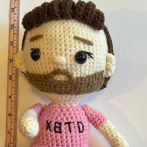 May include: Crocheted doll wearing a pink jersey with the text "XBTD" and the number "10" on it. The doll is holding a white crocheted soccer ball and has brown hair, a brown beard, and black eyes.