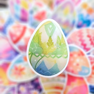Peut inclure: Un autocollant d'œuf en forme d'œuf en aquarelle vert, jaune et bleu avec un contour blanc. L'œuf a un motif de feuilles et une scène de montagne.
