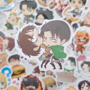 May include: A die-cut vinyl sticker featuring a chibi-style illustration of two characters from Attack on Titan. One character with dark hair and a green cape is holding another character with brown hair. The background is a collage of stickers.