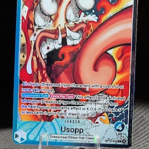 Könnte beinhalten: Sammelkarte mit Usopp von der Dressrosa/Straw Hat Crew. Die Karte zeigt eine lebendige Illustration von Usopp mit einer Stärke von 5000 und Text, der seine Fähigkeiten beschreibt. Die Karte wird auf einem transparenten Kunststoffständer präsentiert.