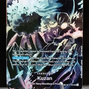 Puede incluir: Una tarjeta coleccionable de One Piece con el personaje Kuzan. La tarjeta tiene un diseño púrpura y azul con el número 5 y 5000. El texto describe las habilidades de la tarjeta. La tarjeta se sostiene en un soporte de plástico transparente.