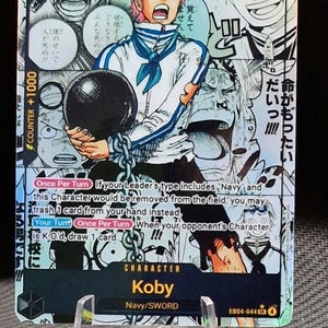 Puede incluir: Una carta de personaje de Koby del juego de cartas One Piece. La carta presenta una ilustración de Koby con un uniforme blanco y azul, sosteniendo una bola y una cadena negras. La carta tiene texto japonés y un poder de ataque de 7000.