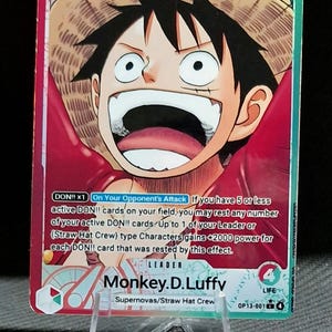 Peut inclure: Carte de collection One Piece avec Monkey D. Luffy, chef de l'équipage du Chapeau de Paille. La carte présente Luffy avec un chapeau de paille, avec une puissance de frappe de 5000. La carte est présentée sur un support transparent.